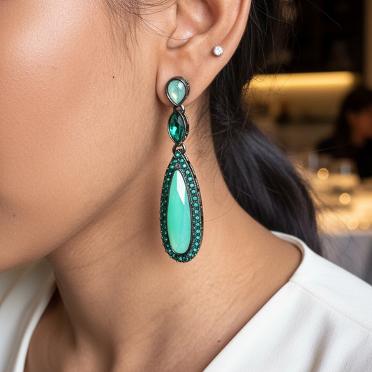 Close-up of a person wearing green teardrop earrings with a blurred indoor background.