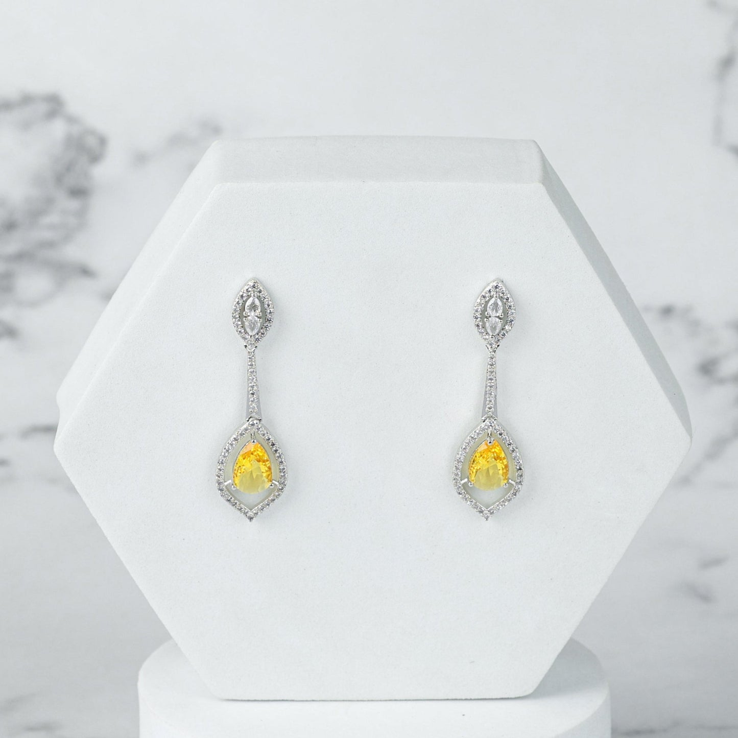 Pair of silver diamond earrings with yellow gemstones on a white stand against a marble background