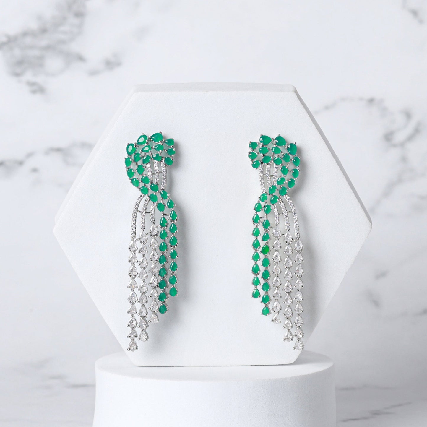 Pair of green and silver diamond earrings on a white pedestal against a marble background