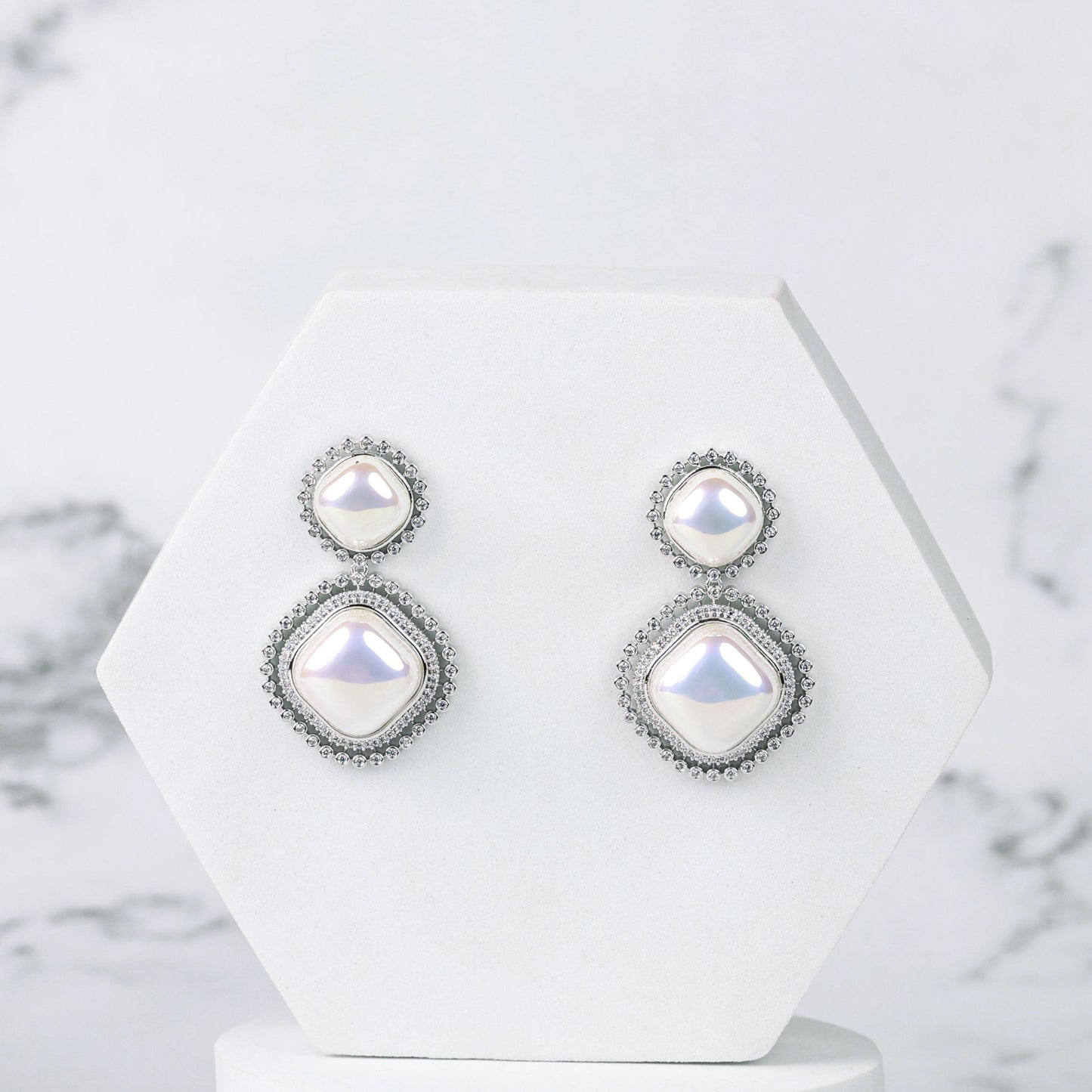 Pair of silver earrings with diamonds and pearls on a white geometric stand against a marble background