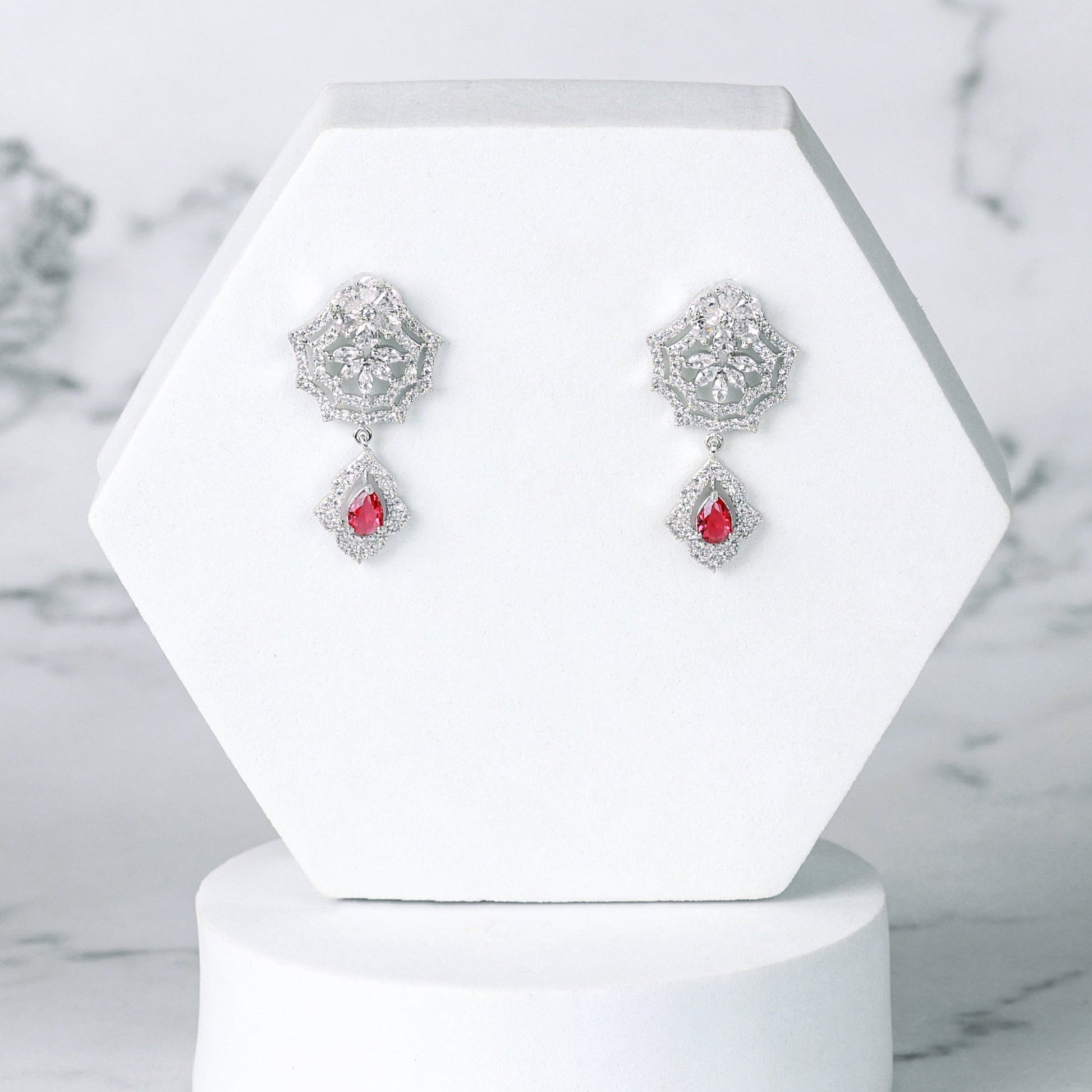 Pair of silver diamond earrings with red stones on a white hexagonal stand against a marble background
