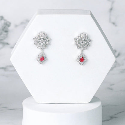 Pair of silver diamond earrings with red stones on a white hexagonal stand against a marble background