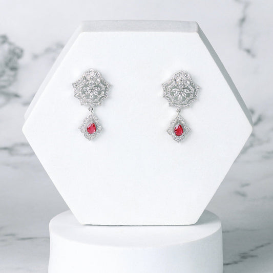 Pair of silver diamond earrings with red stones on a white hexagonal stand against a marble background