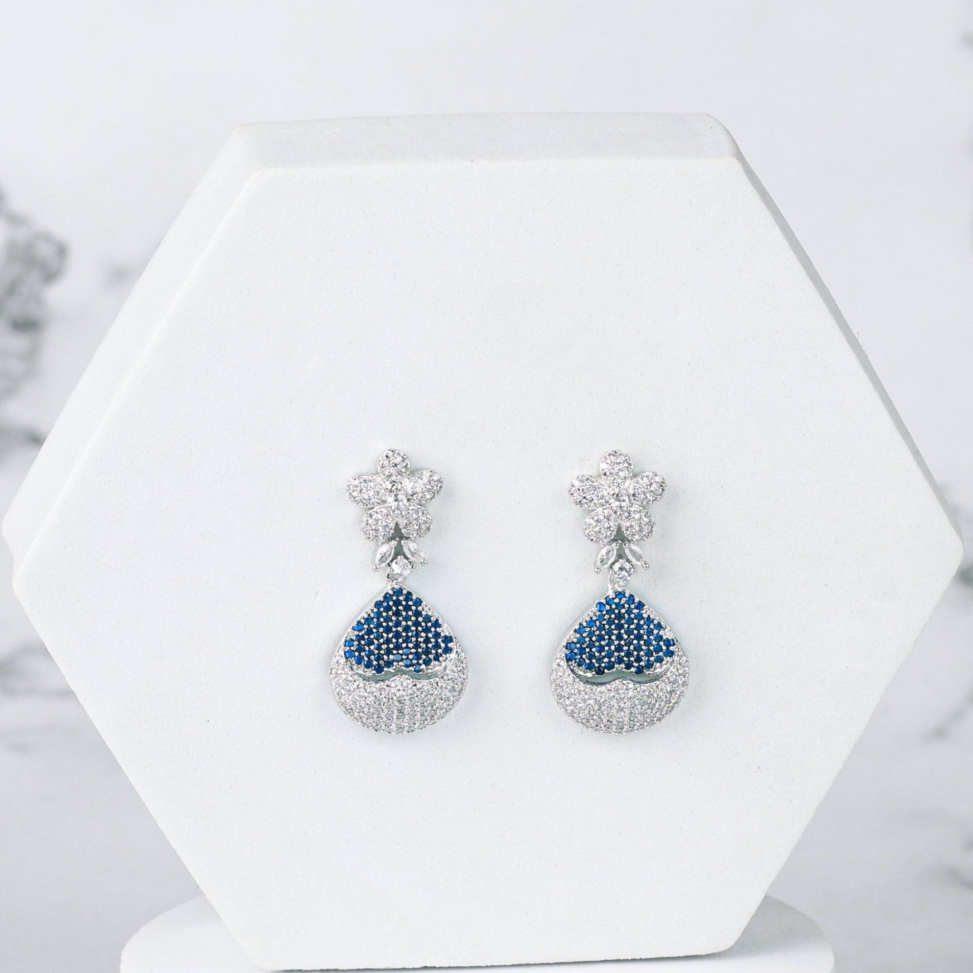 Pair of blue diamond earrings on a white hexagonal stand against a marble background