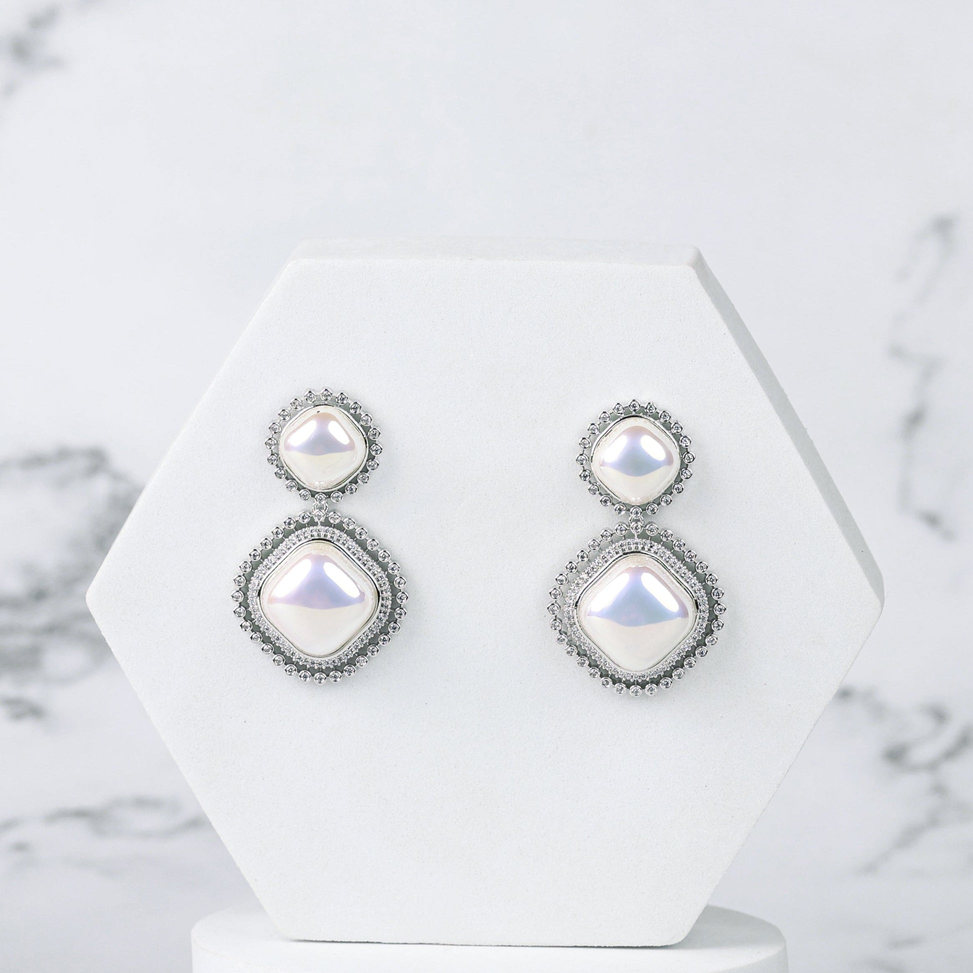 Pair of silver earrings with diamonds and pearls on a white geometric stand against a marble background